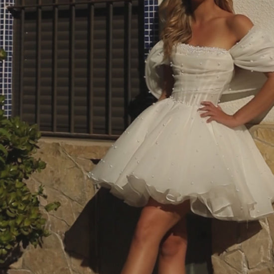 Mini A-line organza wedding dress with off-shoulder draped sleeves, pearl embellishments, and draped bodice. Perfect short reception bridal gown.