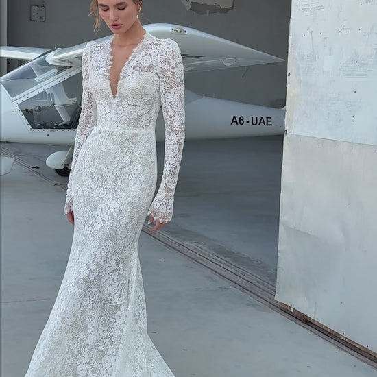 Bride wearing long-sleeve lace fit-and-flare wedding dress with scalloped neckline and chapel train, photographed beside an airplane hangar.