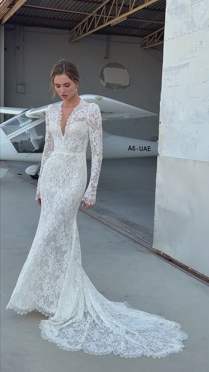 Bride wearing long-sleeve lace fit-and-flare wedding dress with scalloped neckline and chapel train, photographed beside an airplane hangar.