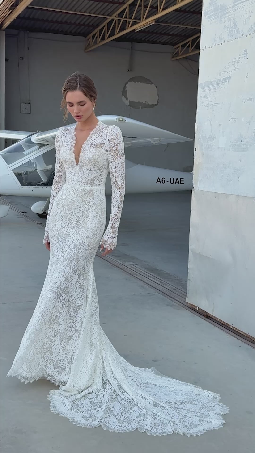 Bride wearing long-sleeve lace fit-and-flare wedding dress with scalloped neckline and chapel train, photographed beside an airplane hangar.