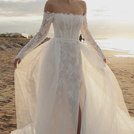 Bride in lace wedding dress with long off-the-shoulder sleeves, slit skirt, detachable tulle overskirt, open boat-shaped back, and sweep court train.