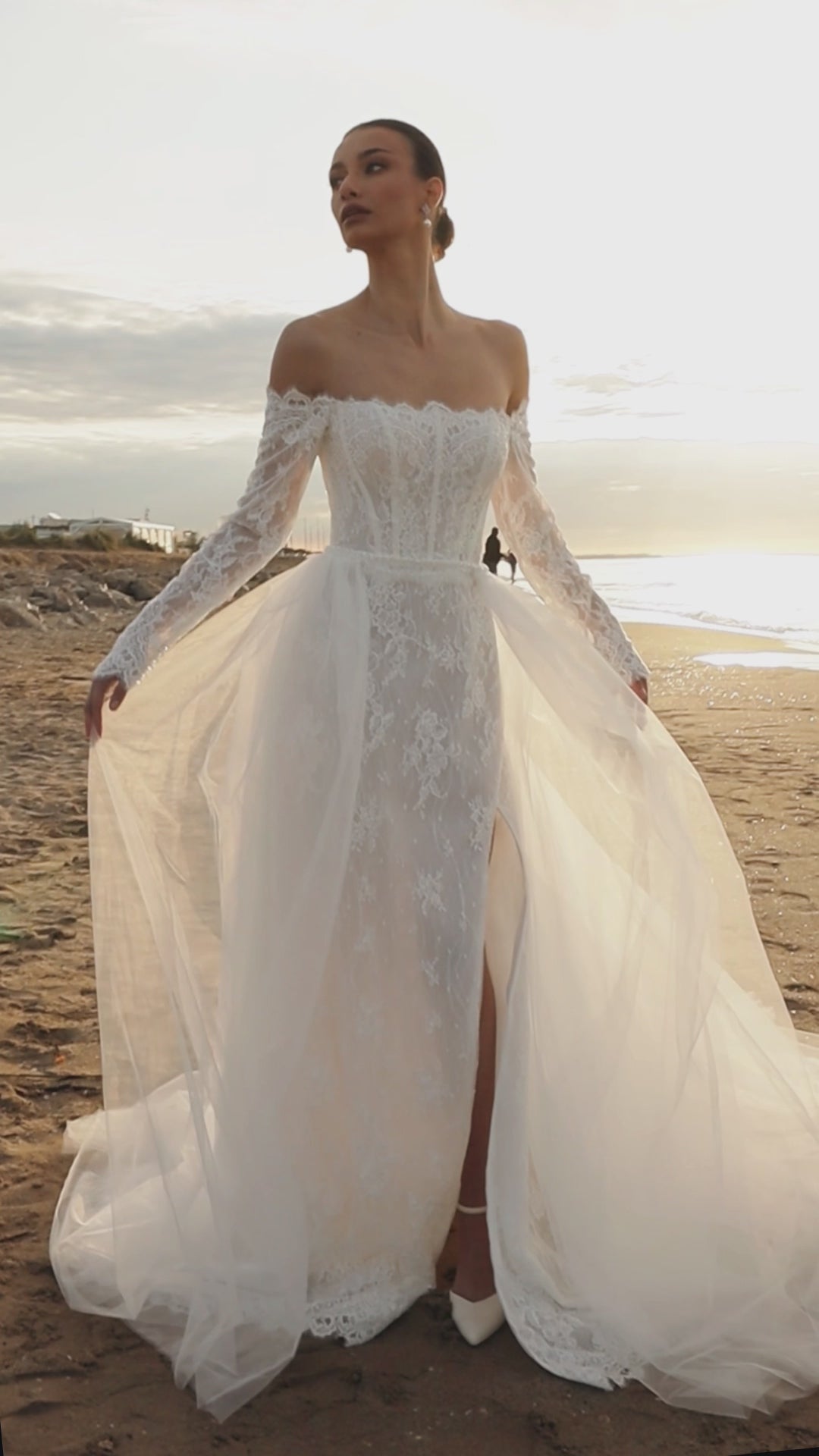 Bride in lace wedding dress with long off-the-shoulder sleeves, slit skirt, detachable tulle overskirt, open boat-shaped back, and sweep court train.