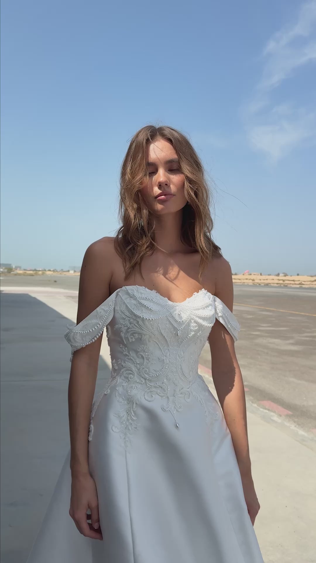 Bride wearing off-the-shoulder Mikado A-line wedding gown with beaded lace bodice and flowing skirt, photographed at an airfield hangar.
