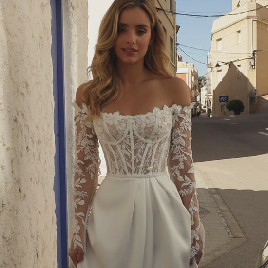 Bride wearing off-the-shoulder boat neckline A-line wedding gown with illusion lace sleeves, lace bodice, slit skirt, zip closure, and court train.