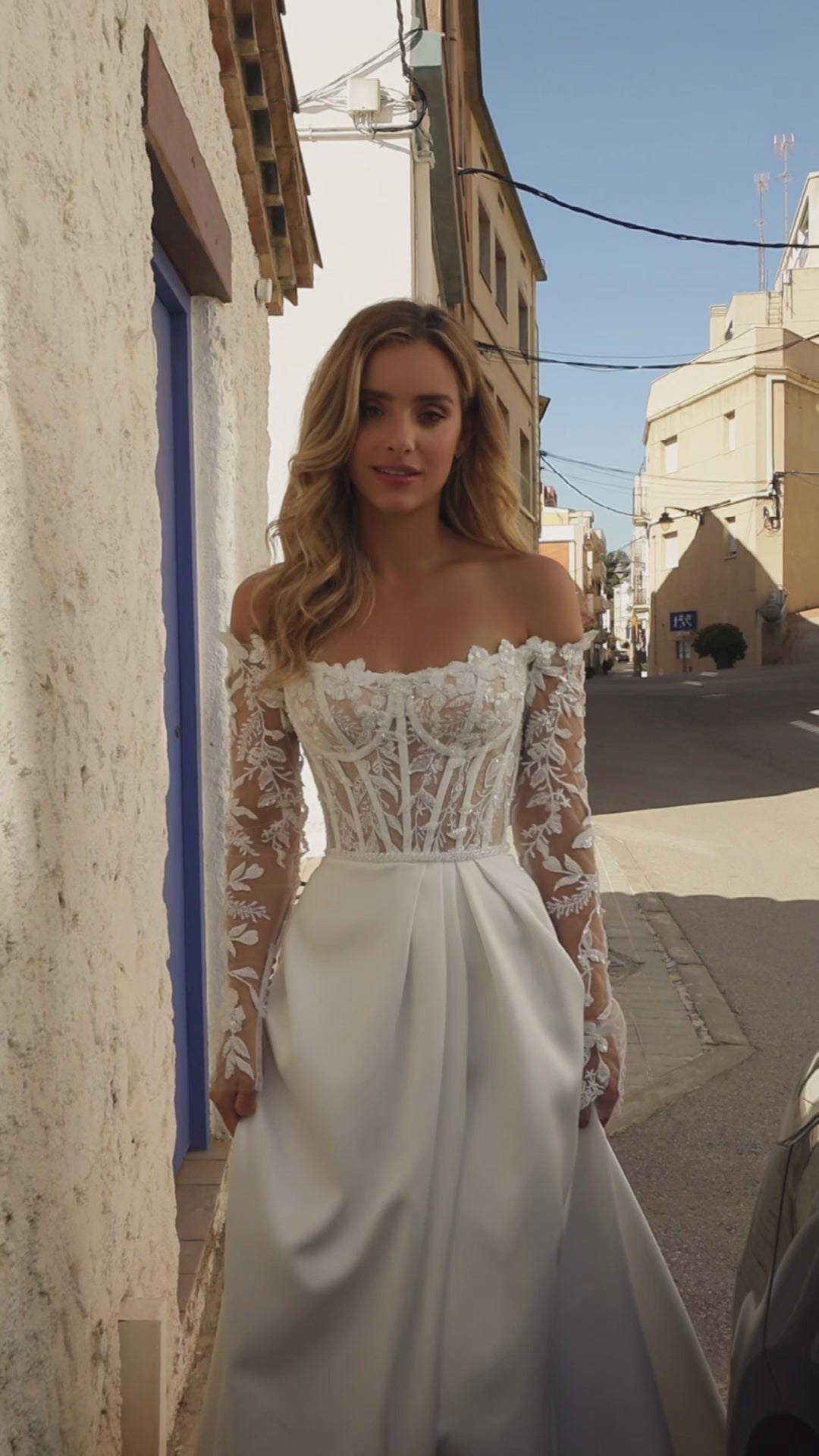 Bride wearing off-the-shoulder boat neckline A-line wedding gown with illusion lace sleeves, lace bodice, slit skirt, zip closure, and court train.