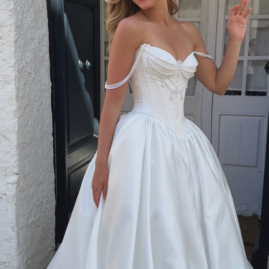 Bride wearing sleeveless satin A-line bridal gown with sweetheart neckline, pearl straps, drop-down waist, zip closure, open back, and chapel train.