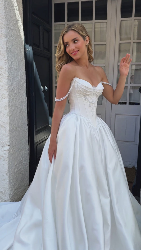 Bride wearing sleeveless satin A-line bridal gown with sweetheart neckline, pearl straps, drop-down waist, zip closure, open back, and chapel train.