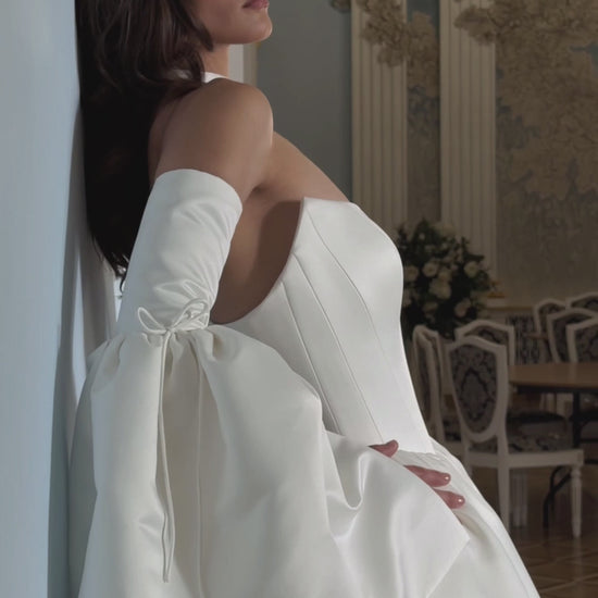Video of an ivory basque waist wedding dress with a structured strapless bodice, detachable bell sleeves, bow choker and flowing skirt.
