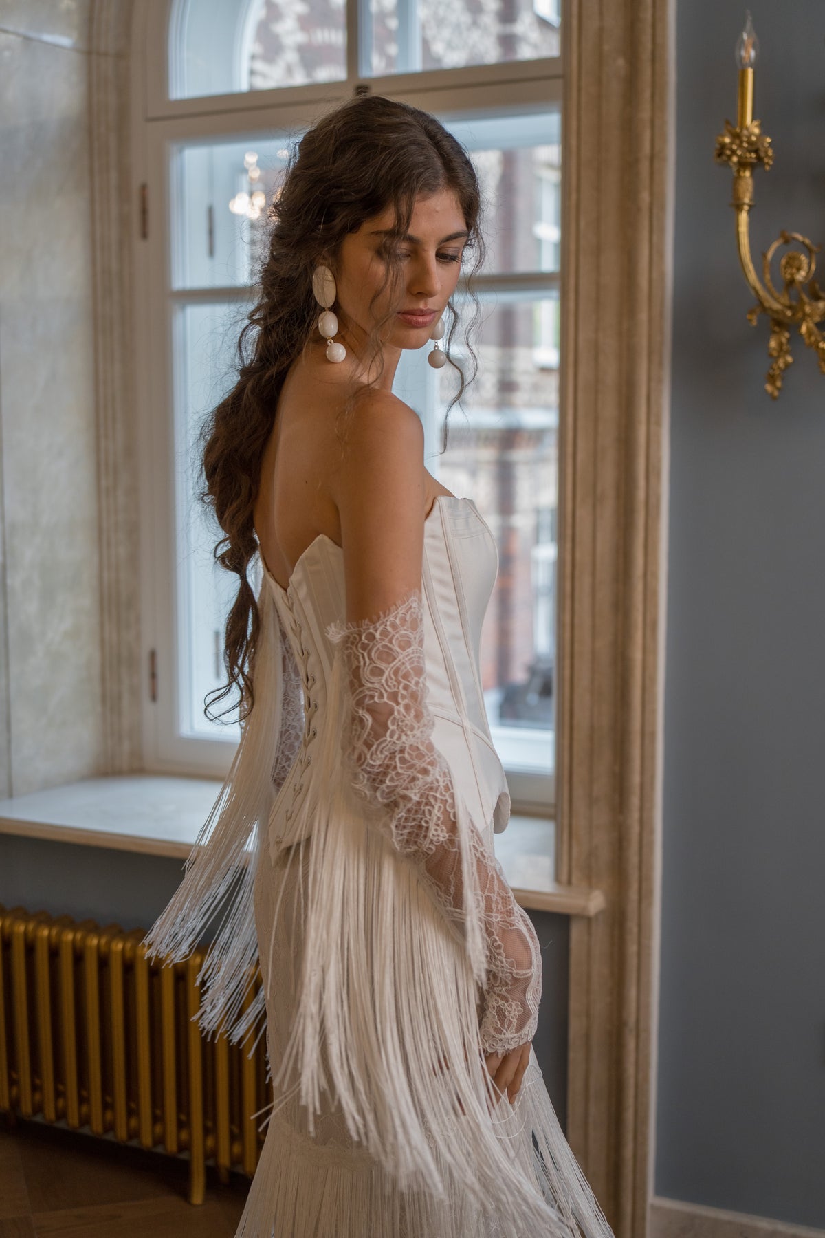 Fitted strapless mikado corset wedding dress with fringe skirt and lace sleeves – side view by a window