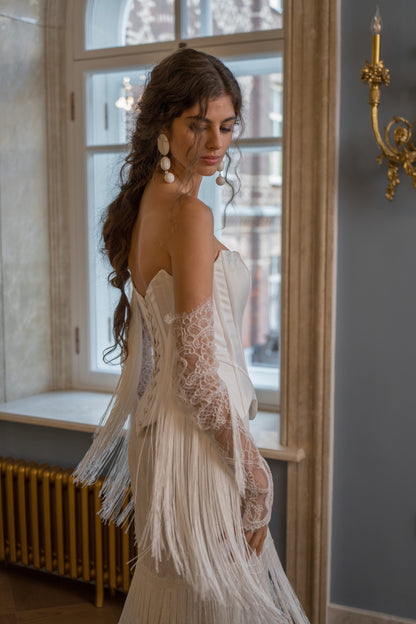 Fitted strapless mikado corset wedding dress with fringe skirt and lace sleeves – side view by a window