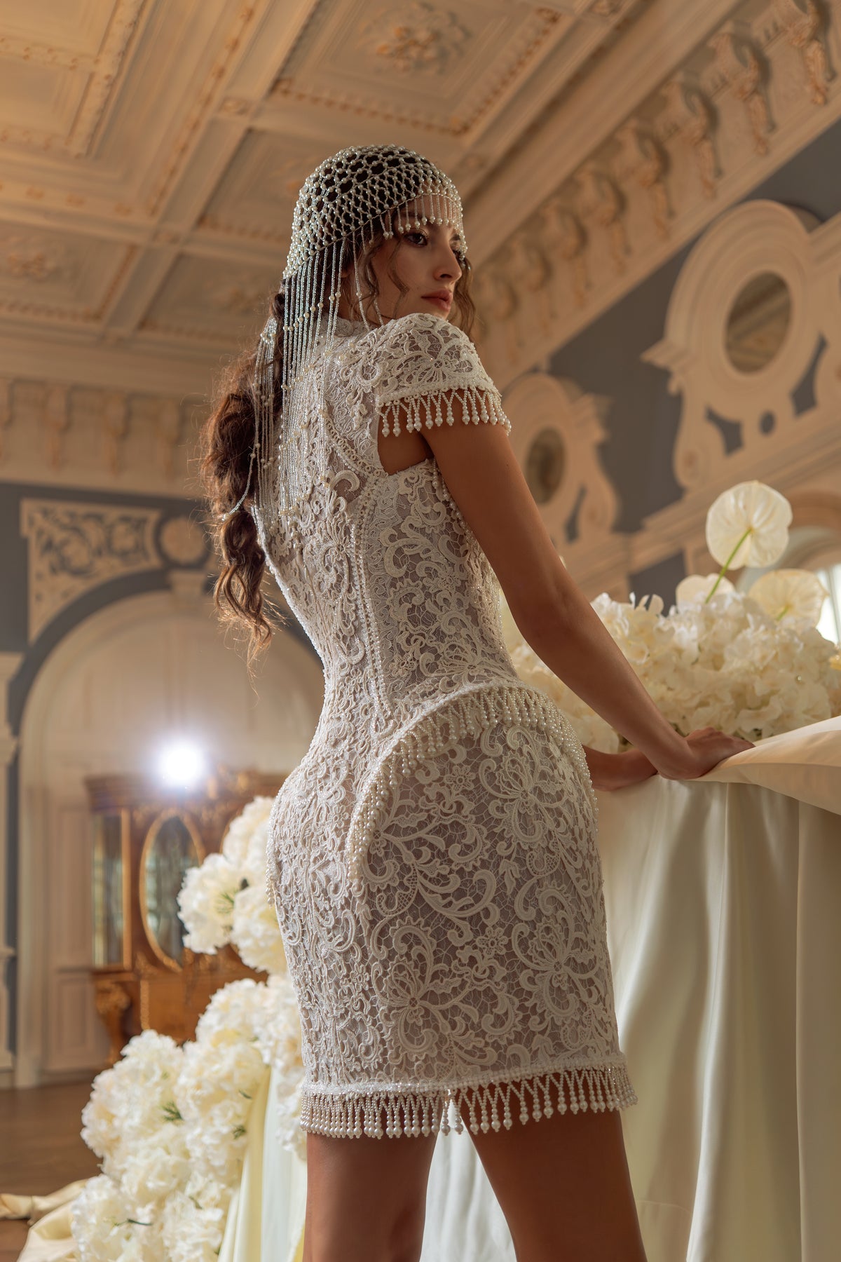 Lace mini wedding dress with pearl fringe – side view leaning by a table with white flowers